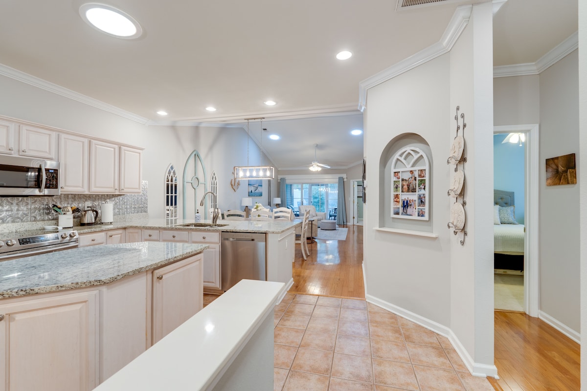 A spacious kitchen features light cabinetry and a large granite countertop, complemented by stainless steel appliances. Natural light enters through a window, creating a bright atmosphere. The kitchen opens to a cozy dining area, with a view of the living area in the background.