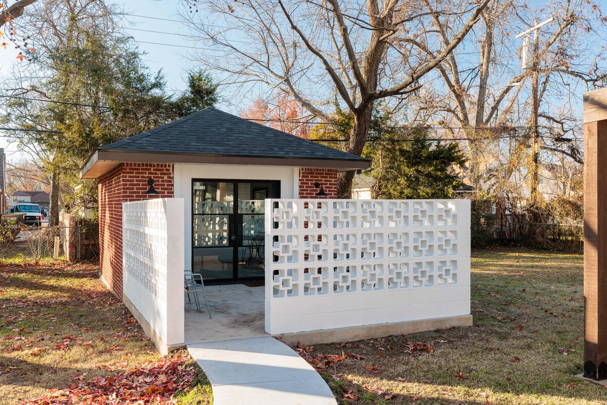 A charming guesthouse is shown, featuring a distinctive brick exterior and a unique decorative block wall. A pathway leads to the entrance, surrounded by well-maintained grass and trees. The sloped roof and large glass doors provide an inviting connection to the outdoor environment.