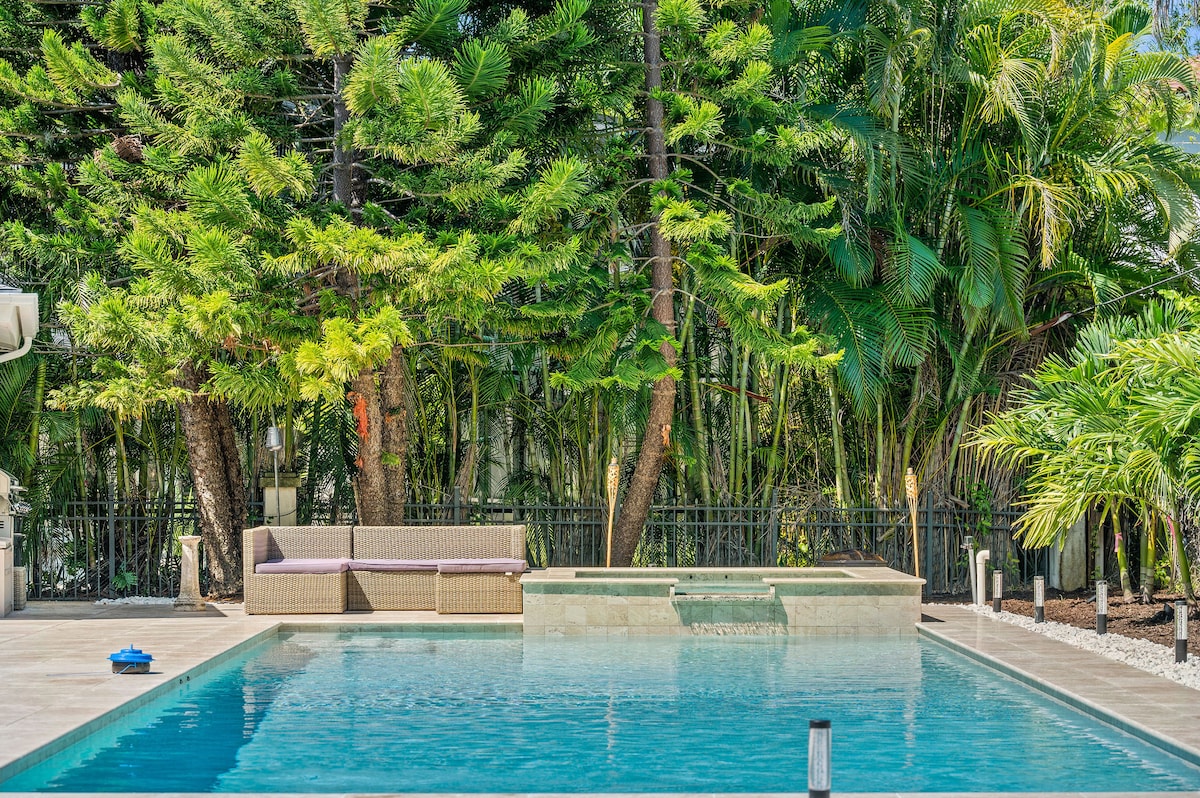 A serene outdoor area features a newly renovated salt swimming pool accompanied by a jacuzzi. Lush greenery and tropical plants surround the space, offering privacy. A comfortable seating arrangement is positioned beside the pool, enhancing the outdoor experience.