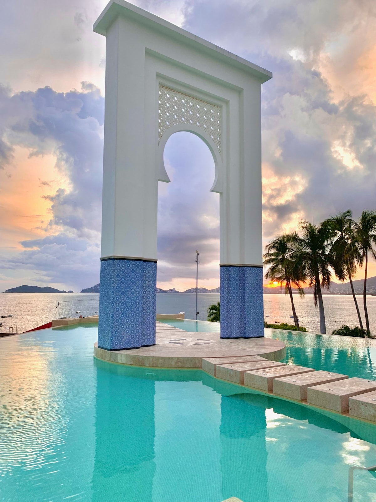 An architectural feature with an arched opening is positioned near a tranquil pool, reflecting the colors of a sunset. Palm trees are seen in the foreground, while distant hills rise over calm waters, enhancing the serene coastal atmosphere.