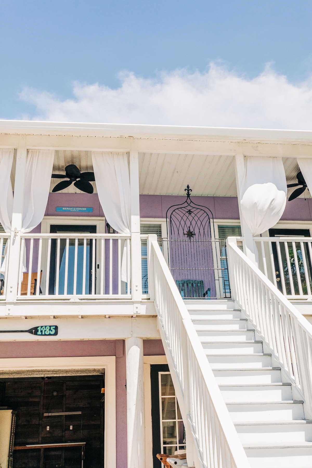 The exterior of the beach house features a two-story design with a staircase leading to the upper level. Light, sheer curtains drape from the balcony, complementing the lavender-hued walls. Ceiling fans are positioned on the porch, providing a breezy atmosphere.