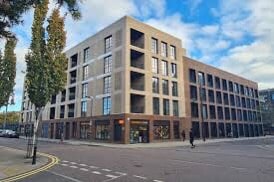 The modern building features a mix of brick and glass facades with large windows, allowing for plenty of natural light. Several ground-floor shops are visible, providing convenience to residents. Trees line the street, contributing to a welcoming urban environment.