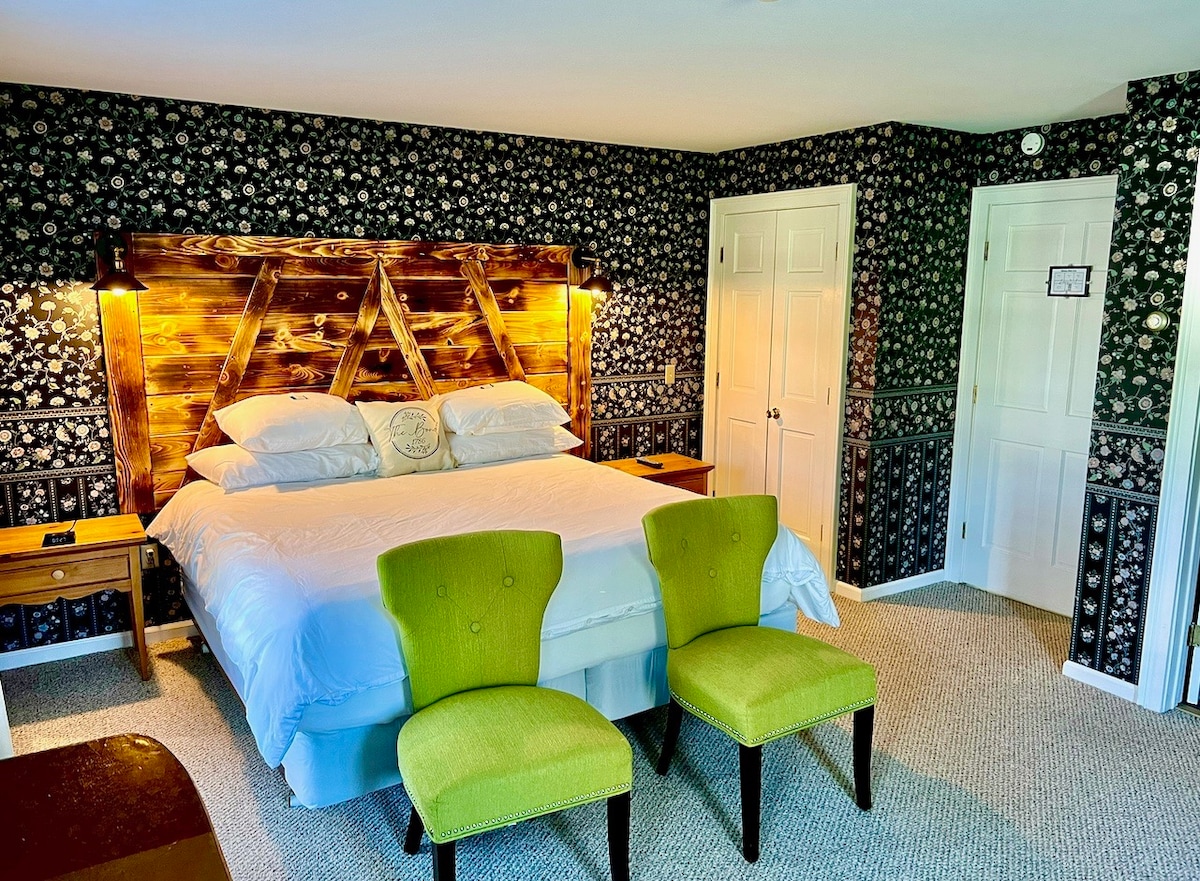 A cozy bedroom features a spacious bed with crisp white linens and a unique wooden headboard illuminated by soft lighting. Two green chairs are positioned at the foot of the bed. The walls are adorned with patterned wallpaper, enhancing the room's charm.
