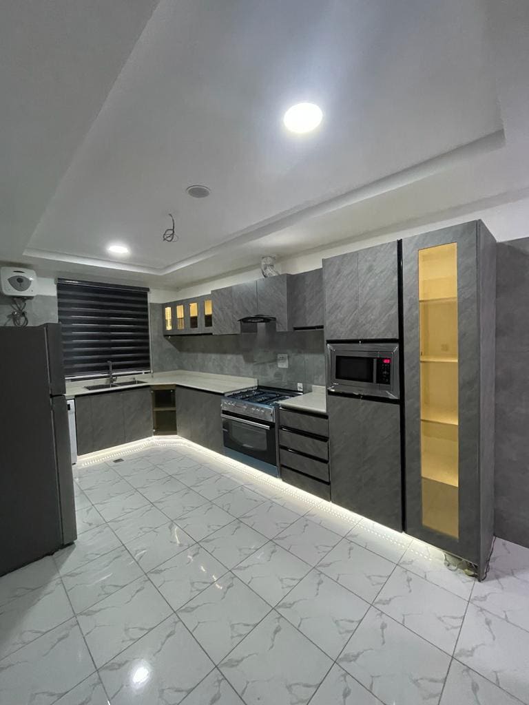 A modern kitchen features sleek grey cabinetry with gold accents, complemented by stainless steel appliances. An elegant countertop offers ample workspace, while under-cabinet lighting enhances visibility. The spacious layout includes a refrigerator, oven, and a microwave, all arranged for convenience.