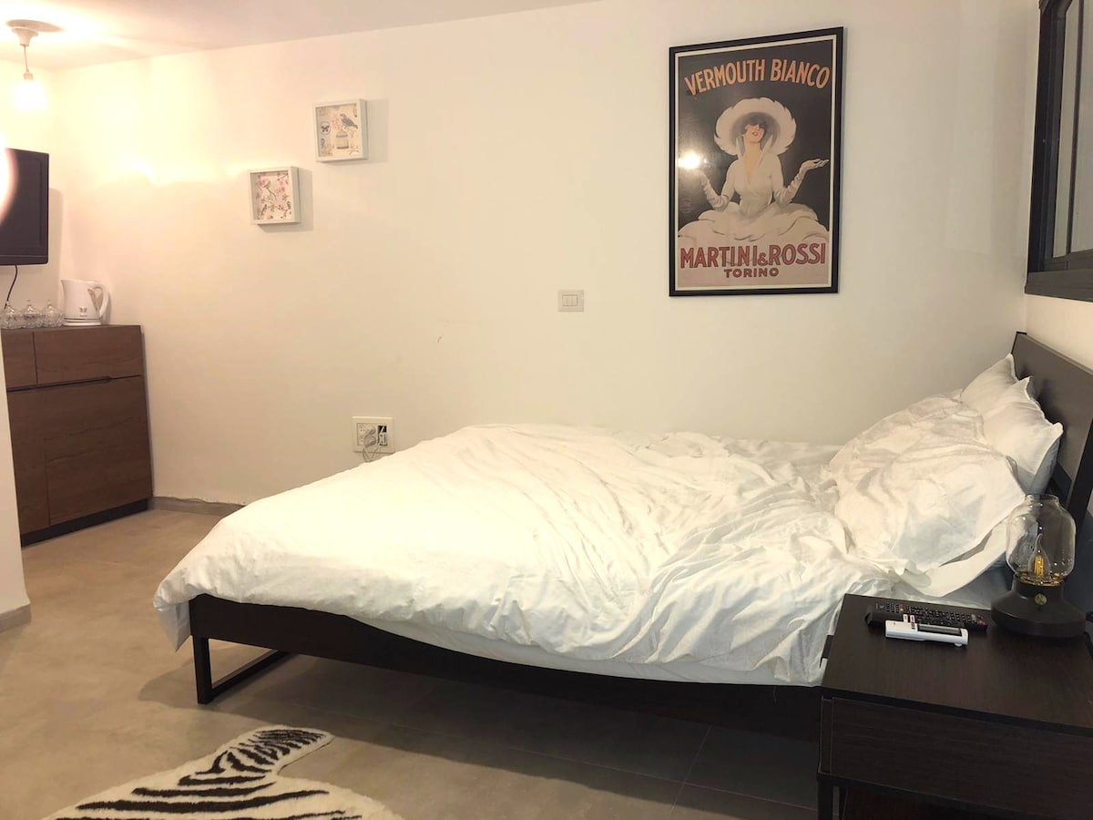 A well-lit bedroom contains a spacious bed topped with white linens. A wooden nightstand and a dresser are present, along with framed wall art and a television. The room features a neutral color palette and a tiled floor for a clean, modern look.
