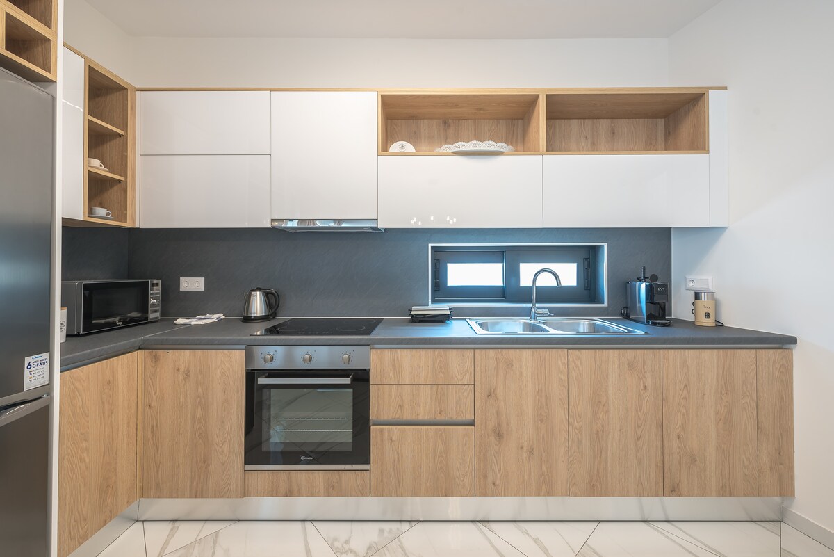 The modern kitchen features sleek cabinetry with a blend of wood and white finishes. Appliances include a stove, oven, and microwave, complemented by a stainless steel kettle. Ample storage space is provided by open shelves displaying dishes and kitchen essentials. The countertop design incorporates subtle textures.
