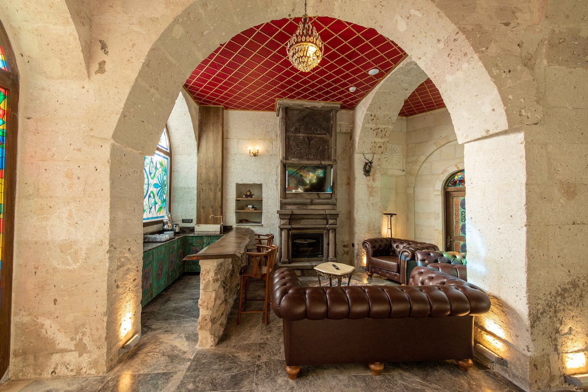 A spacious lounge area is highlighted by stone archways and a red ceiling. A brown leather sofa is positioned invitingly, near a traditional fireplace. Natural light filters through stained glass windows, illuminating the space. A wooden bar area is visible in the background.