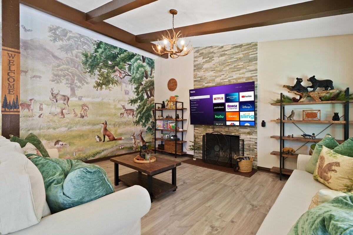 A cozy living room features a nature-themed mural depicting deer and a serene landscape. The space includes two soft sofas with green throw pillows, a coffee table at the center, and a wall-mounted smart TV. Rustic decor adds to the inviting atmosphere.