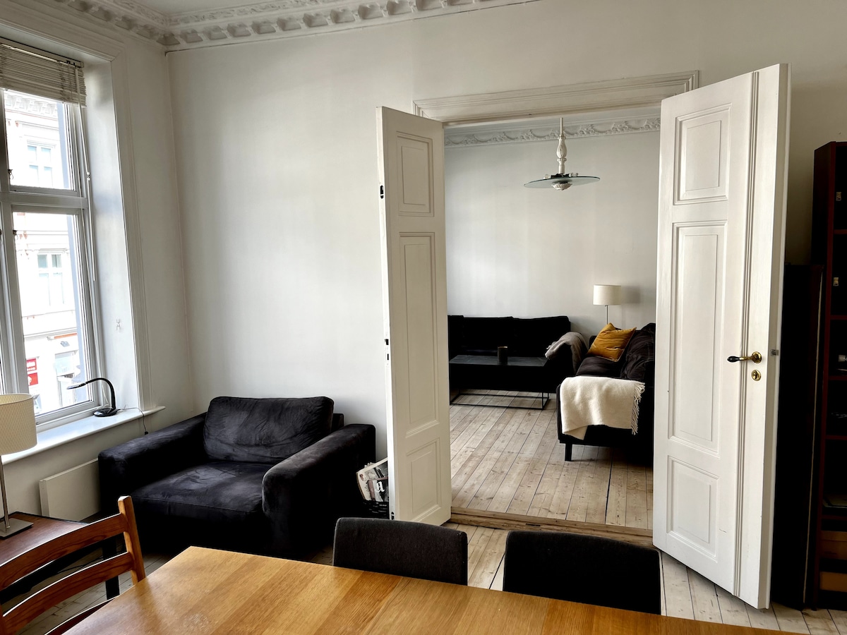 An open doorway reveals a spacious living area featuring a comfortable black sofa and a light-colored throw. Wooden floors and high ceilings enhance the sense of airiness. A dining table with chairs is positioned in the foreground, offering a welcoming space for meals.