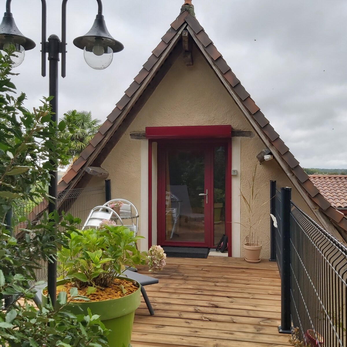 A charming outdoor balcony is featured, showcasing wooden decking and a small table with chairs. Lush greenery and decorative plants surround the space. The entrance to the room, highlighted by a bright red overhang, offers direct access, enhancing the connection to nature.