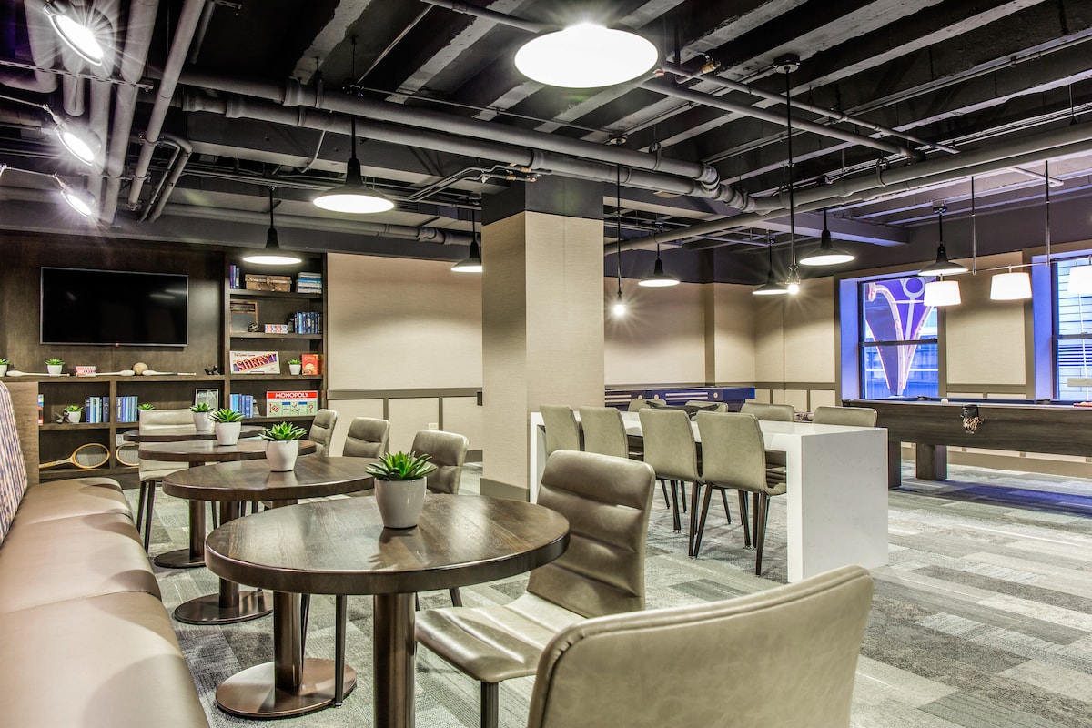 A modern communal space is highlighted by several round wooden tables and comfortable seating. Soft lighting illuminates the area, where a large television and shelves filled with books are visible. Large windows allow natural light to fill the room, creating a bright atmosphere.