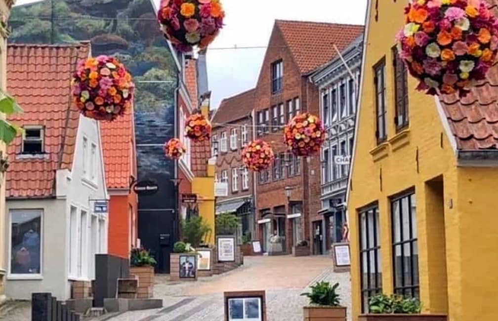 A charming street scene features two rows of colorful buildings with red-tiled roofs. Hanging floral arrangements add vibrancy above the cobblestone path. The area is lined with planters and shops, creating an inviting environment for exploration.