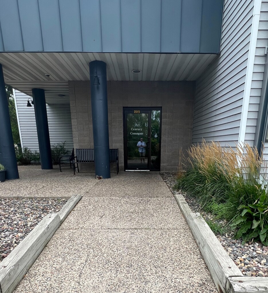 The entrance to the apartment building is seen, featuring a glass door flanked by two tall columns. A pathway leads to the door, bordered by gravel and low greenery. A bench is positioned to the left, offering a place to sit.