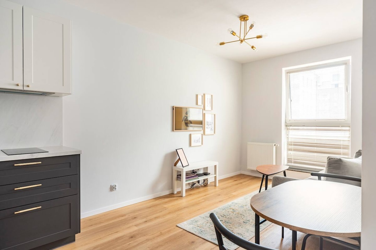 A modern living space features light wood flooring and soft grey walls. A small dining table with black chairs sits beside a compact side table. Wall art decorates the room, complemented by a large window allowing natural light to fill the area.