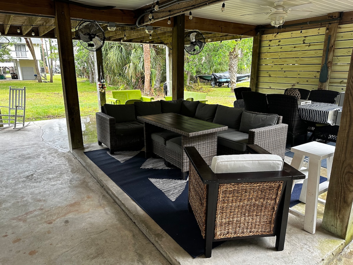 A covered outdoor seating area is presented with comfortable wicker furniture, featuring a rectangular table at the center surrounded by L-shaped and single chairs. A large area rug adds texture underfoot, while ceiling fans and open views of the lawn enhance the atmosphere.