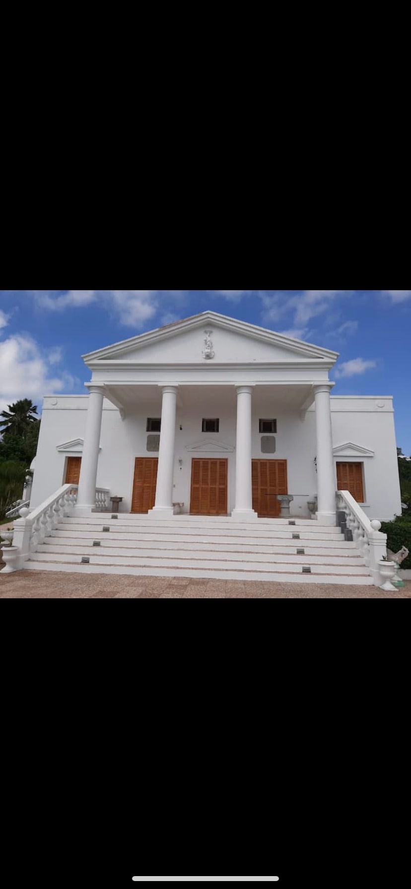 A grand white building features a symmetrical façade with four tall columns supporting a gabled roof. Wooden doors are positioned on both levels, flanked by elegant staircases. The clear blue sky complements the striking architecture.