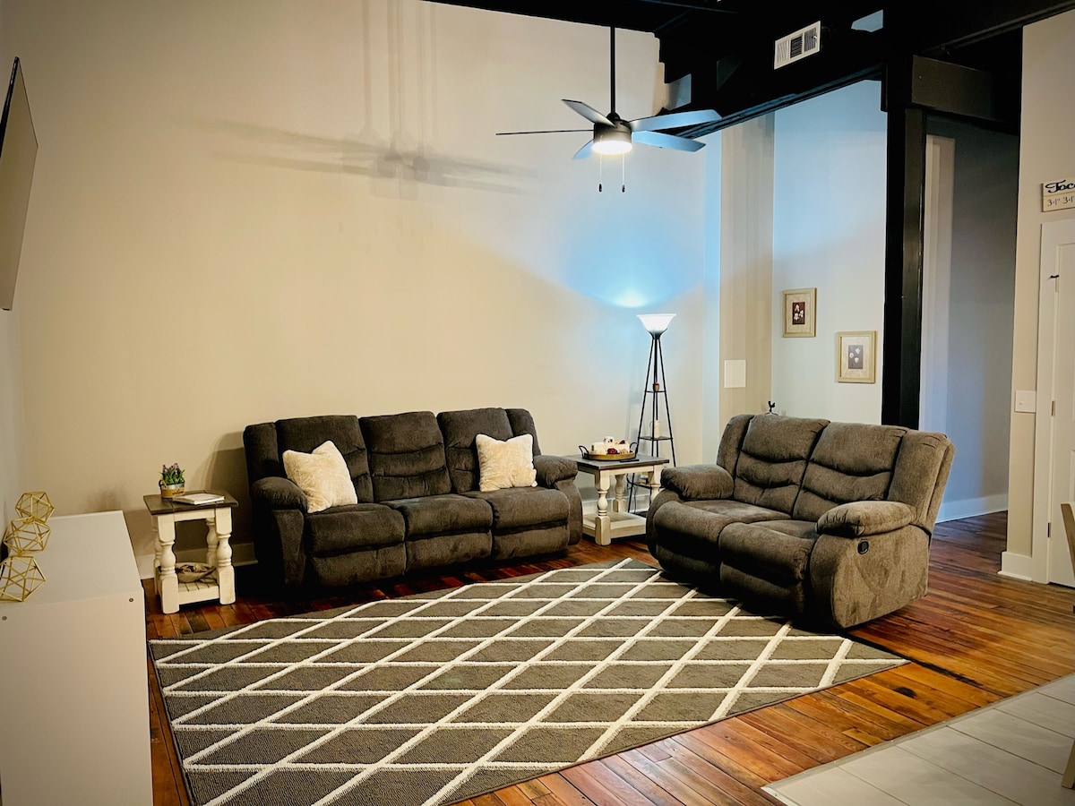 A spacious living area is featured with two comfortable grey sofas arranged around a soft area rug with a geometric pattern. A floor lamp provides additional lighting, and wall decor enhances the modern feel of the space.