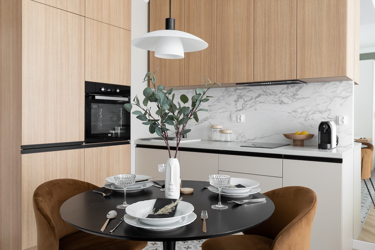 A modern kitchen area features wooden cabinetry and sleek marble backsplash. A round black table is set with elegant dishware and cutlery, while a small vase with greenery adds a natural touch. A coffee maker is visible on the countertop.