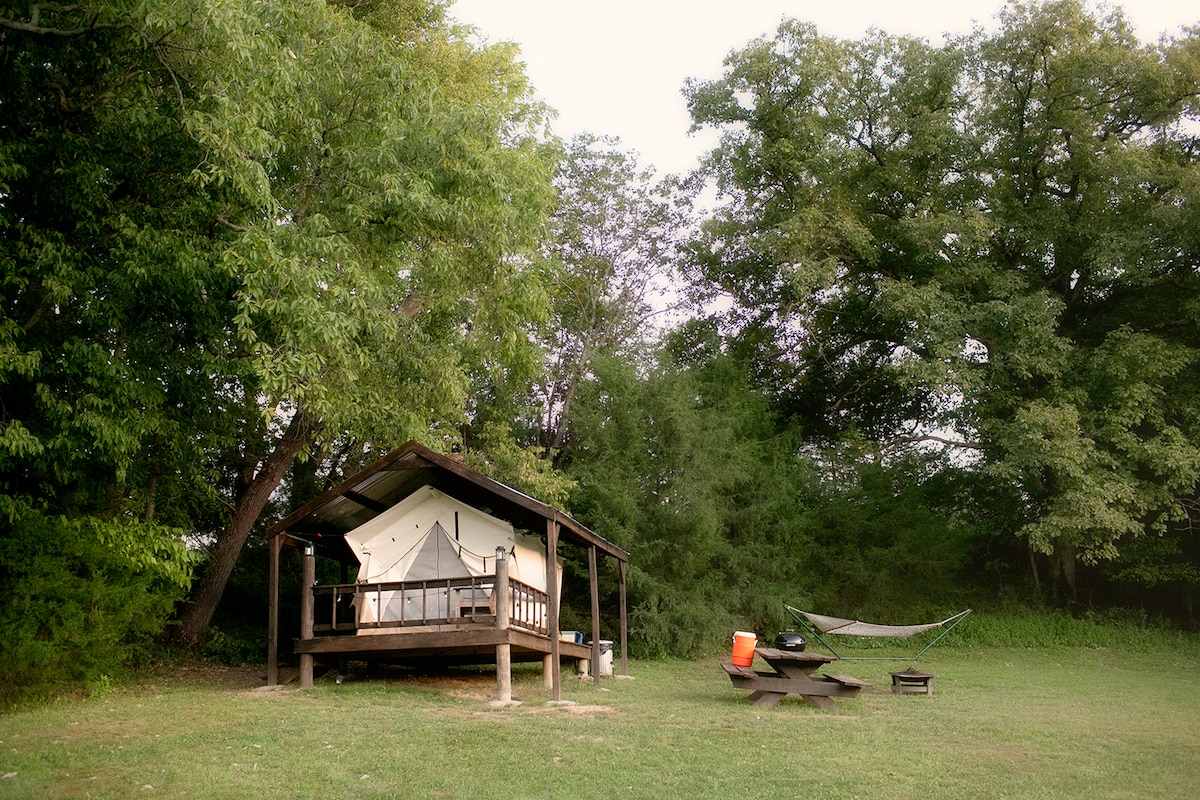 A canvas tent is situated on a raised deck, surrounded by greenery. Nearby, a hammock is hung between trees, and a picnic table is positioned on the grass. An orange cooler is visible, adding to the campsite atmosphere.