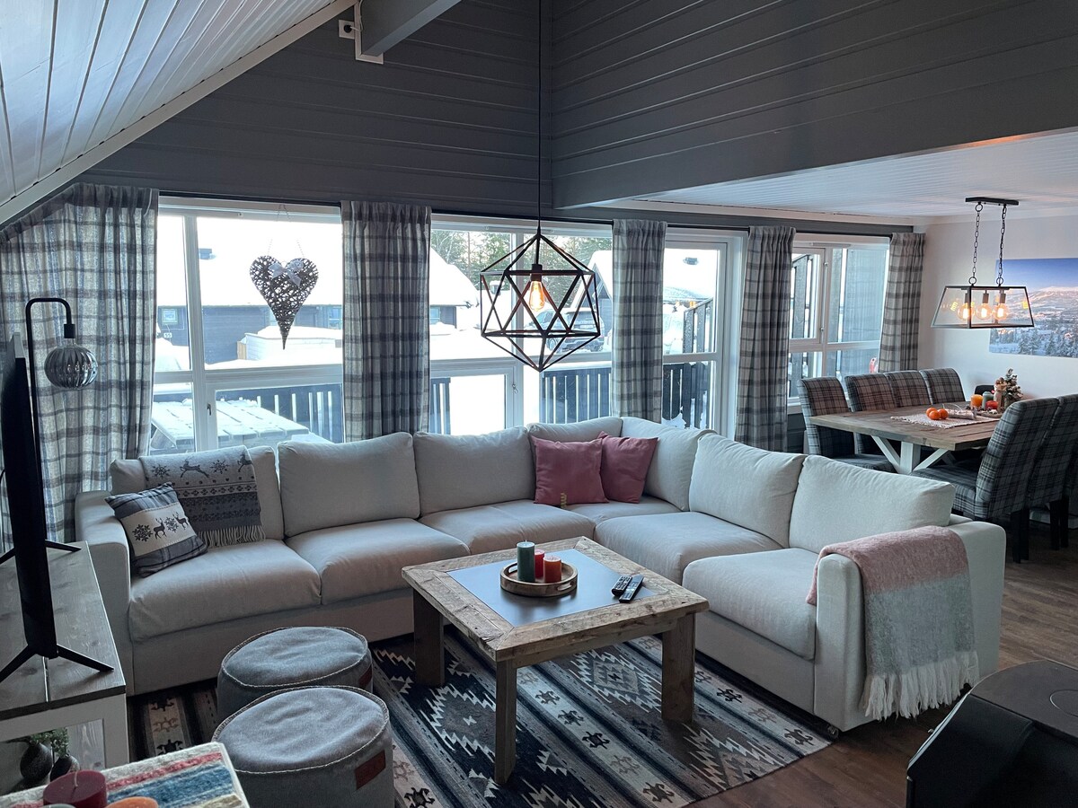 A spacious living area is featured with a large, L-shaped sofa adorned with a pink cushion and a patterned throw. A wooden coffee table sits at the center. Large windows provide ample natural light, framed by plaid curtains, with views of the snowy outdoor landscape.