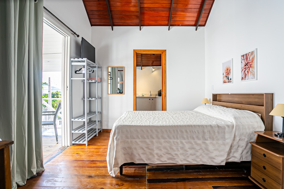 A bright sleeping area features a spacious bed with light bedding, complemented by bedside tables and lamps. Natural light enters through an open door leading to a balcony. A mirror and an open kitchen area can be seen in the background, enhancing the functional layout.