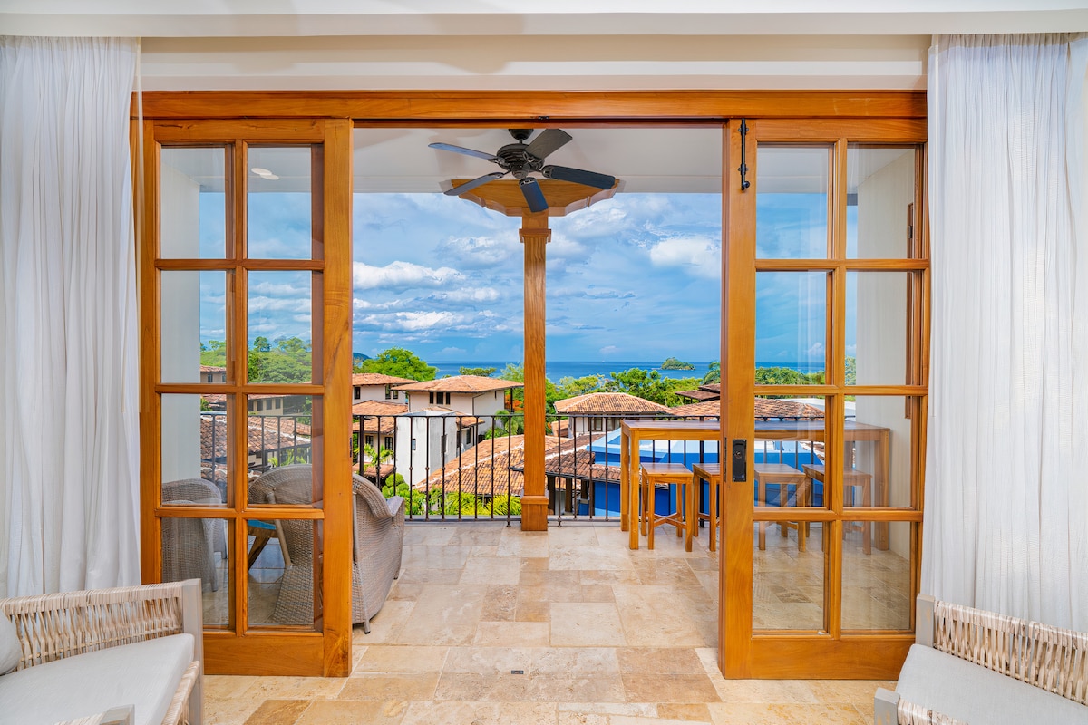 Glass doors provide access to a spacious balcony with ocean views. Comfortable outdoor seating is arranged nearby, alongside a dining table. Light-colored curtains frame the view, enhancing the connection to the natural surroundings and inviting the ocean breeze.