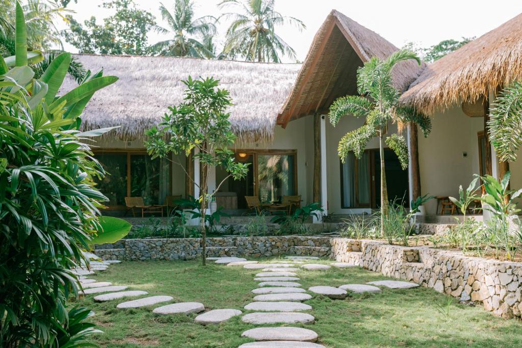 A serene outdoor space is shown, featuring a pathway of stone steps leading through a lush garden. Thatched-roof structures are visible in the background, surrounded by tropical plants and palm trees, offering a sense of tranquility and connection to nature.