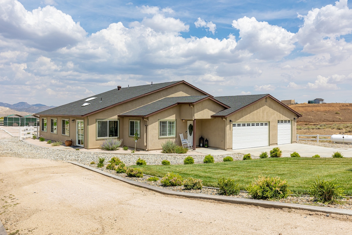 The exterior of a modern single-story home is presented, with a neutral-colored façade and a welcoming driveway. Lush green landscaping and shrubs complement the surroundings, while large windows allow natural light. The spacious property is set against a backdrop of mountains under a partly cloudy sky.