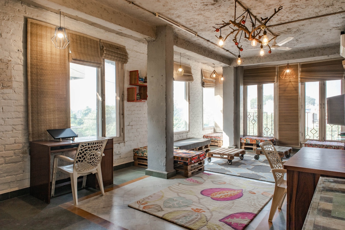 A bright common room features large windows that allow natural light to fill the space. Colorful seating is arranged around a wooden coffee table on a patterned rug. A small desk is positioned in one corner, complemented by decorative light fixtures hanging from the ceiling.