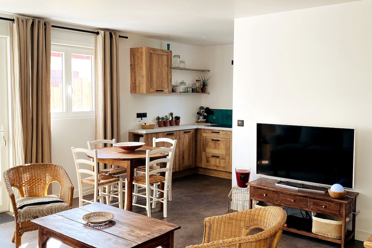 The interior living area features a wooden dining table surrounded by white and natural woven chairs. A compact kitchen with wooden cabinetry and teal backsplash is visible in the background. A flat-screen TV is positioned on the wall opposite a comfortable seating arrangement.
