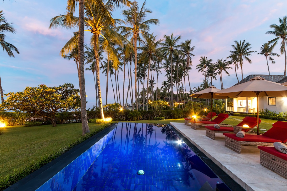 An outdoor space features a vibrant blue infinity pool surrounded by lush greenery and tall palm trees. Comfortable lounge chairs with red cushions are positioned beside the pool. Soft lighting creates an inviting atmosphere as the sun sets, casting a warm glow across the scene.