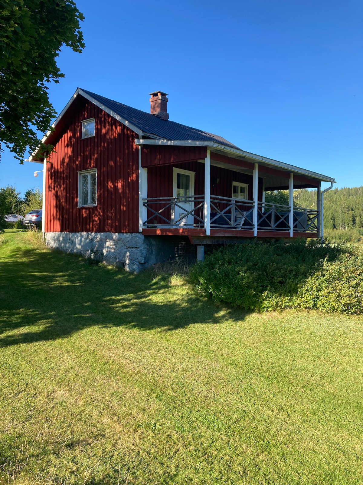 A charming red cabin is positioned on a grassy hillside, featuring a welcoming porch supported by wooden pillars. The landscape is surrounded by trees, creating a peaceful environment. A stone foundation complements the rustic appearance of the structure against a clear blue sky.