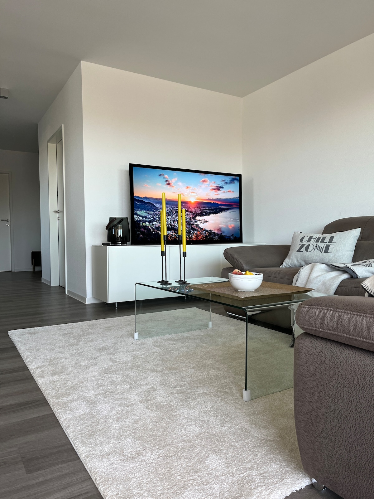 A cozy living area is presented with a plush sofa and a glass coffee table. A digital screen displaying a scenic view is mounted on the wall, while a soft area rug enhances the space. Decorative candles add a touch of warmth.