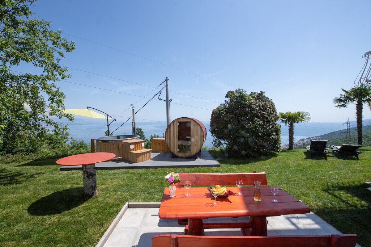 A serene outdoor space is displayed, featuring a red picnic table and a round wooden deck amidst lush green grass. A kitchenette and covered seating area are visible, with panoramic views of the water and distant hills under a clear blue sky.