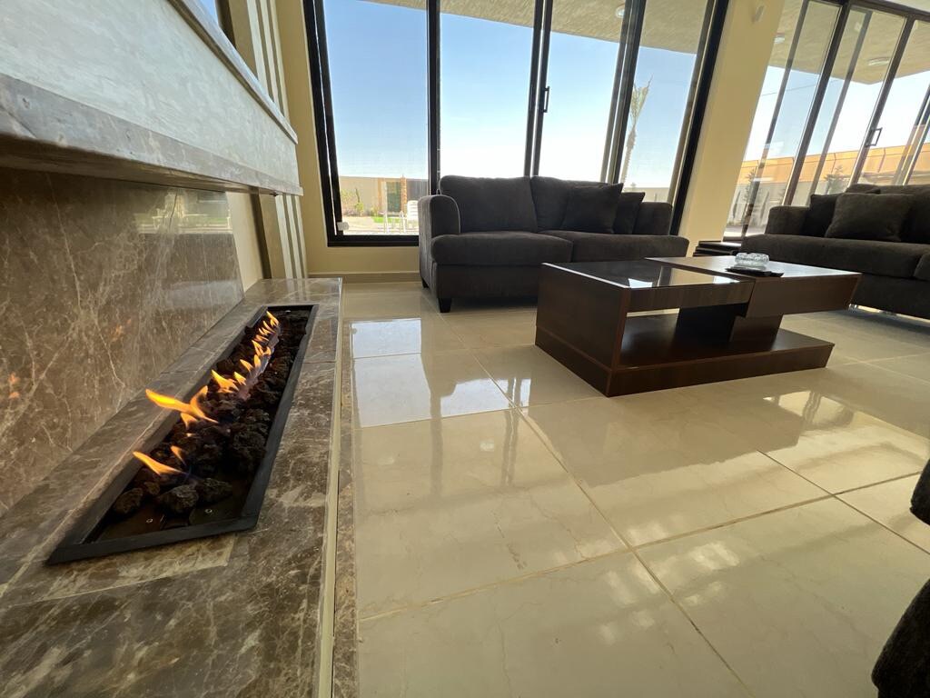 A modern living area features a sleek fireplace with flickering flames set into a marble wall. Two comfortable sofas face a dark wooden coffee table, placed on shiny tiled flooring. Large glass windows allow natural light to fill the space, offering views of the outdoors.
