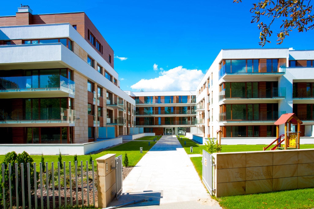 A modern apartment complex is set against a clear blue sky, featuring structured balconies and large windows. Green grass surrounds a central pathway leading to the entrance, with a children's playground visible on the right, enhancing the family-friendly environment.