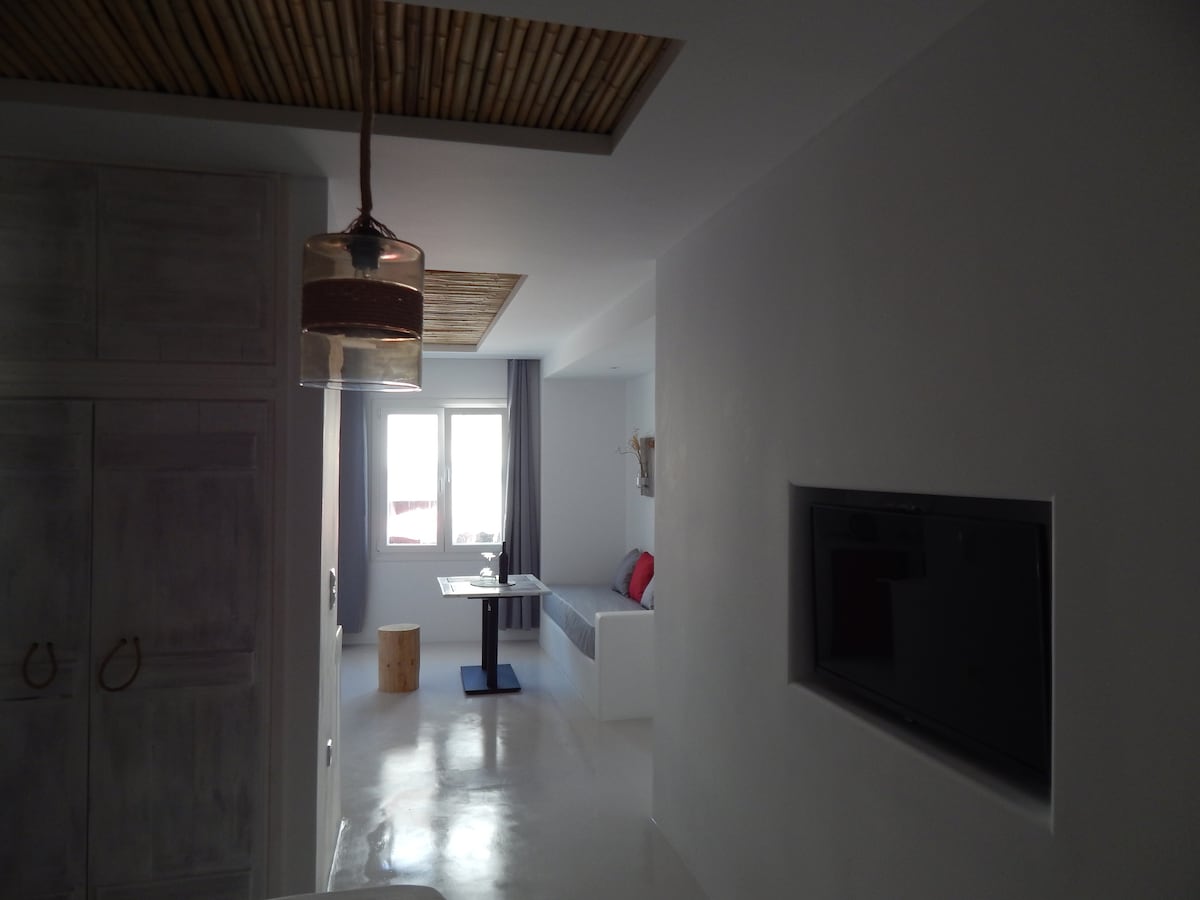 A bright corridor leads to a living area, featuring a comfortable seating arrangement and a small dining table. Natural light fills the space through a window, while a television is mounted on a white wall. Subtle textures are added by the wooden ceiling design.