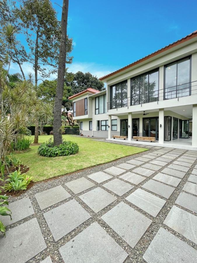 A modern villa is depicted, featuring large windows and a spacious patio that opens to a well-maintained garden. The stone pathway leads through vibrant greenery, while the trees provide a natural shade. The clear sky enhances the peaceful ambiance of the surrounding area.