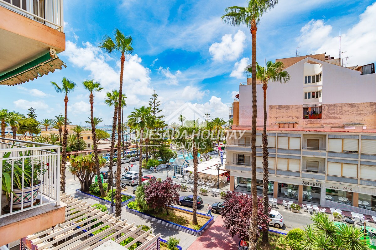 A vibrant street view overlooking palm trees and landscaped greenery. The scene captures a lively atmosphere with nearby restaurants and shops, framed by modern buildings under a bright blue sky with fluffy clouds. Sunlight enhances the welcoming urban environment.