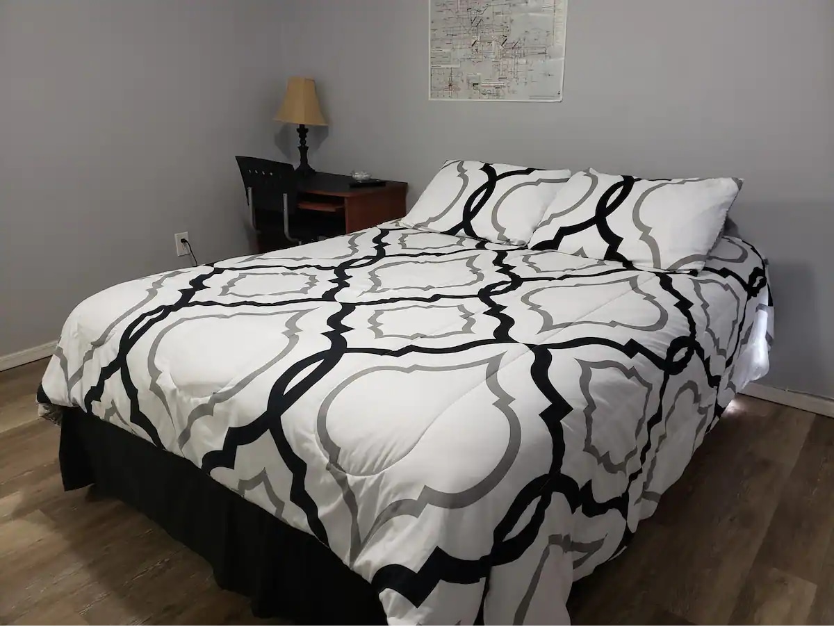 A queen sized bed is positioned against a light grey wall, adorned with a patterned black and white comforter. A small desk with a lamp is located beside the bed, providing a functional workspace. Natural light filters in, enhancing the calming environment.