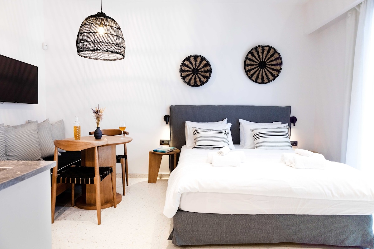 A modern studio apartment features a spacious bed adorned with soft linens. A round dining table is present, accompanied by stylish chairs. Decorative wall hangings add texture to the bright, minimalist space, while a warm light fixture enhances the overall ambience.