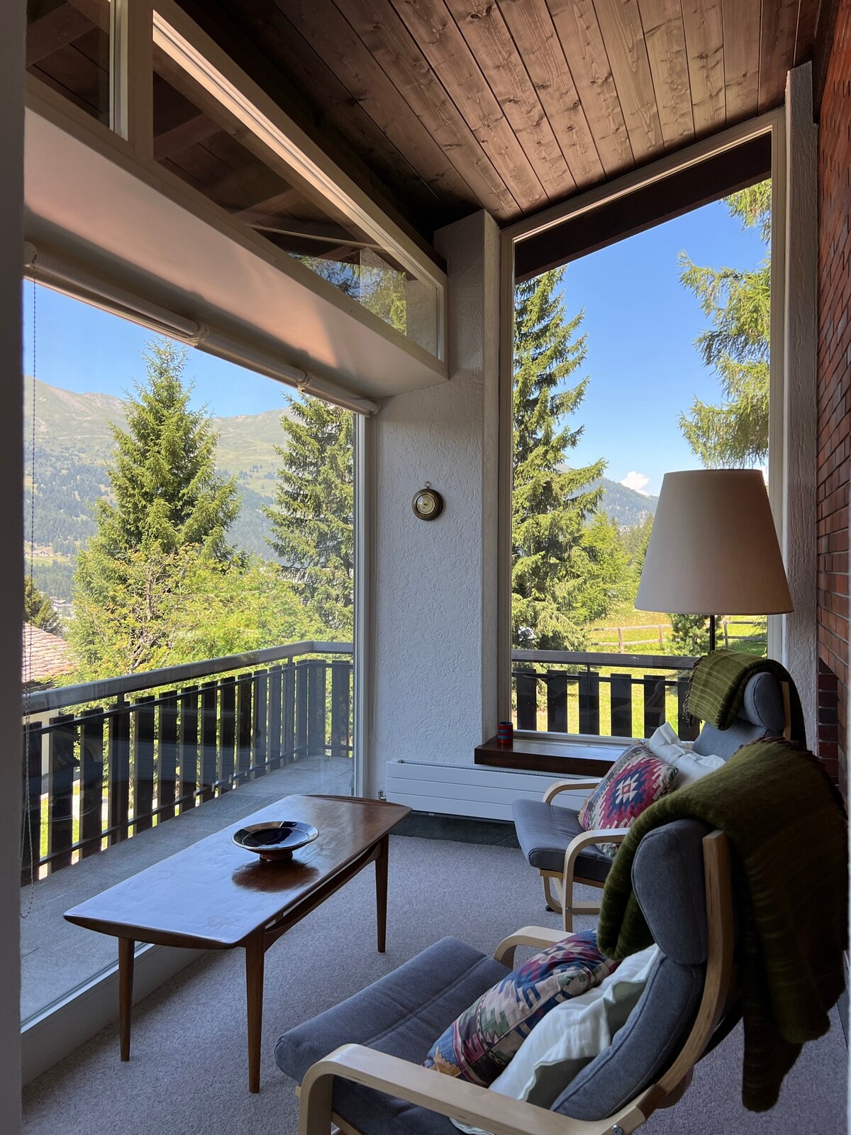 A cozy seating area is situated on a covered balcony, featuring two comfortable chairs and a wooden table. Large windows provide expansive views of surrounding trees and mountain landscape. A warm blanket is draped over one chair, and a decorative bowl rests on the table.