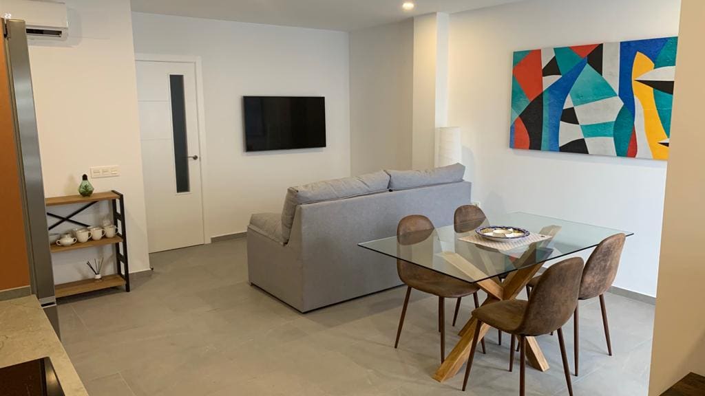 A modern living space features a comfortable gray sofa positioned near a flat-screen television. A glass dining table with wooden legs accommodates several chairs. Bright artwork adorns the white wall, enhancing the contemporary design of the area, filled with natural light.
