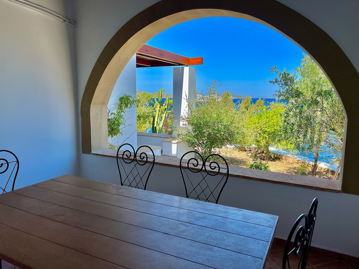 An arched window frames a view of the sea and greenery outside. A wooden dining table with metal chairs occupies the foreground, creating an inviting space. The bright blue sky complements the natural surroundings, enhancing the serene atmosphere within.