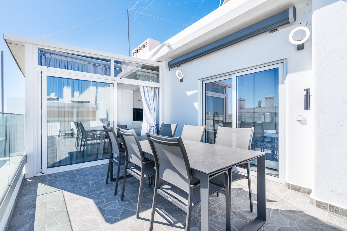 A private terrace equipped with a spacious dining table and six chairs is featured. Glass doors provide views of the surroundings, and large windows allow natural light to fill the space. The terrace overlooks a clear blue sky, enhancing the outdoor ambiance.
