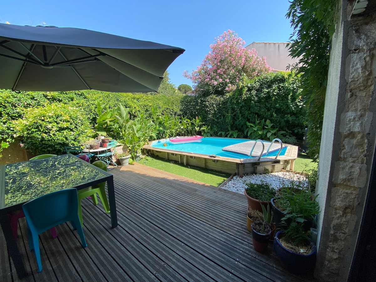 A private outdoor area is depicted, featuring a round pool surrounded by lush greenery. A wooden deck provides space for a table with colorful chairs. A large umbrella offers shade, while potted plants add natural elements to the serene environment.