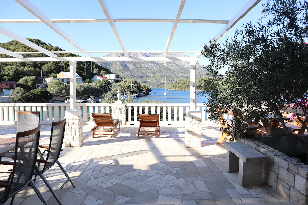 An outdoor terrace features a spacious stone-paved area with wooden lounge chairs positioned towards the water. Framed by a white pergola, the scenic view includes lush hills and a tranquil bay, creating a serene environment for relaxation.