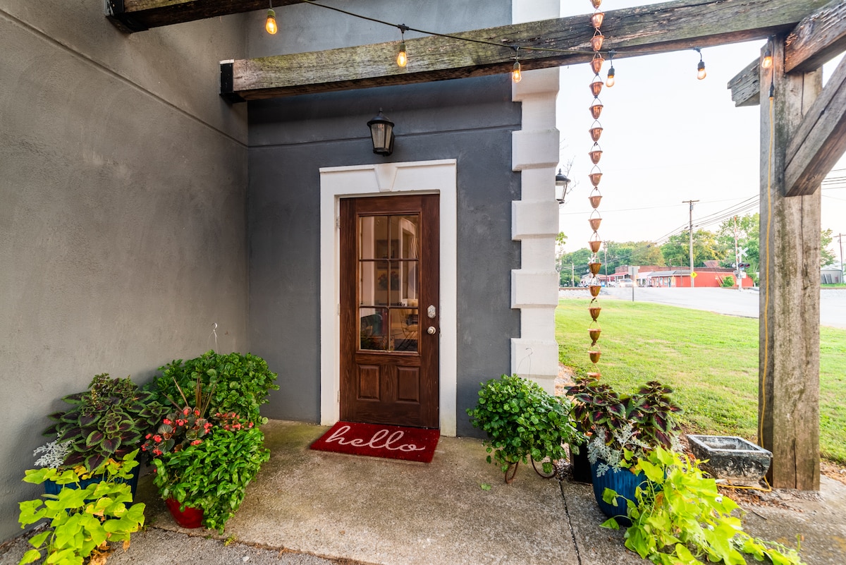 The entrance features a welcoming wooden door bordered by potted plants and a cheerful mat that reads 'hello.' Soft lighting is provided by string lights overhead, enhancing the atmosphere. A grassy area is visible beyond the entrance.