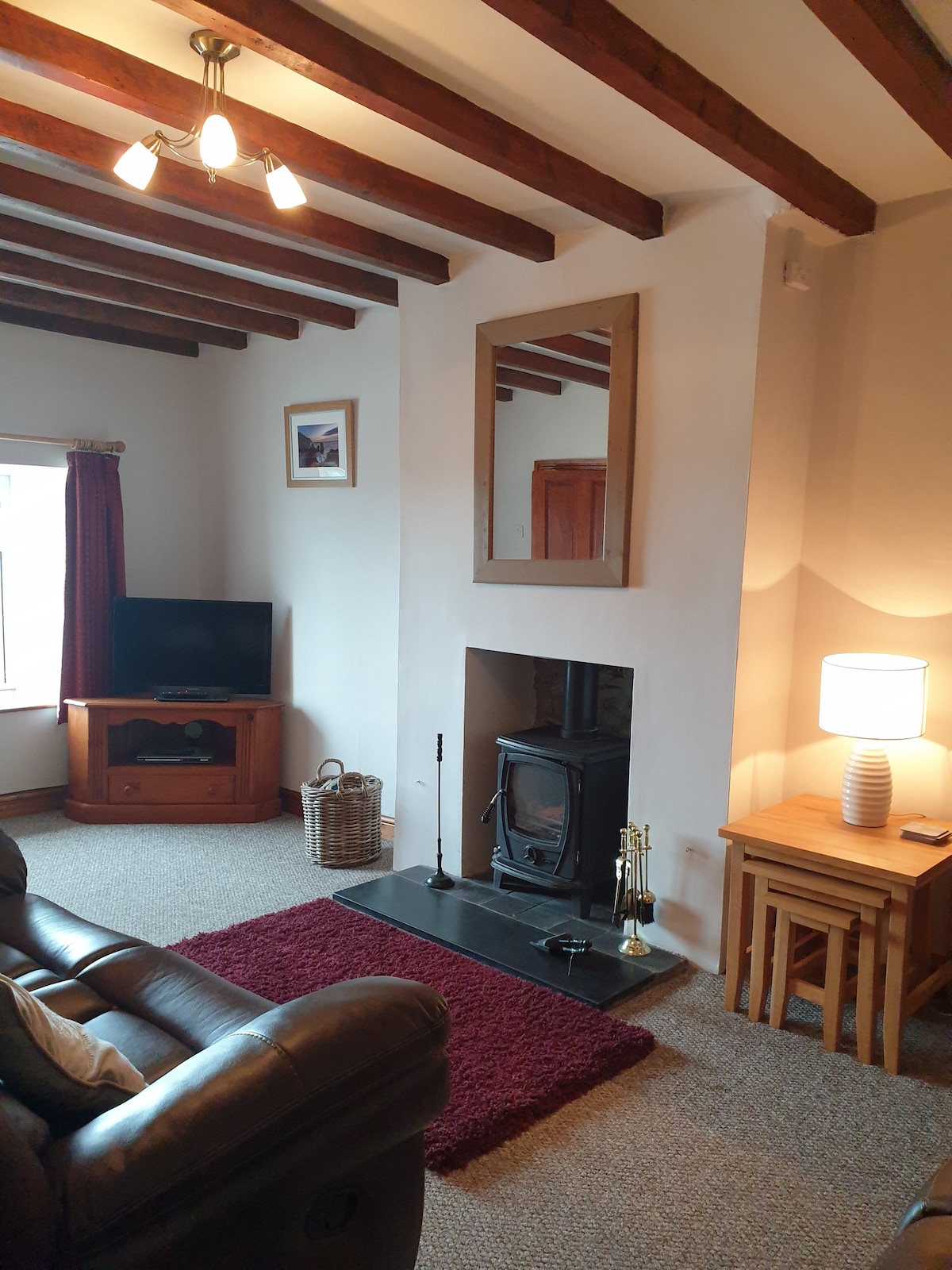 The living room features a beamed ceiling with a cozy log burner, complemented by a comfortable seating area. A television is placed on a wooden cabinet, and a mirror is mounted above the fireplace. A table with a lamp provides soft lighting, enhancing the inviting atmosphere.