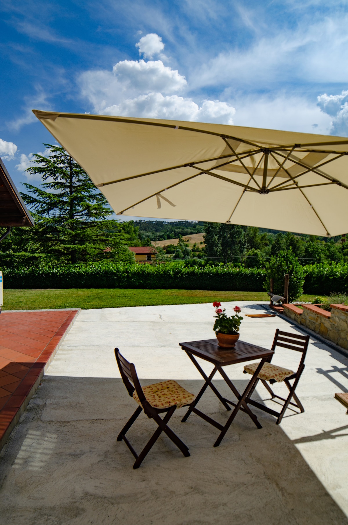 An outdoor seating area features a small wooden table accompanied by two folding chairs. A potted plant sits at the center of the table. A large umbrella provides shade, while a scenic view of rolling hills and greenery can be seen in the background under a bright blue sky.